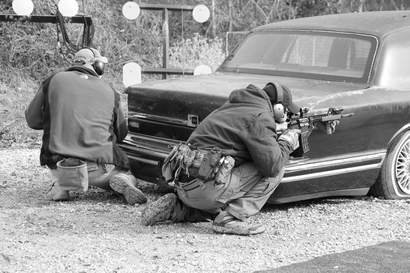 Ballistic team members training behind a car as cover, one aiming a carbine at steel targets