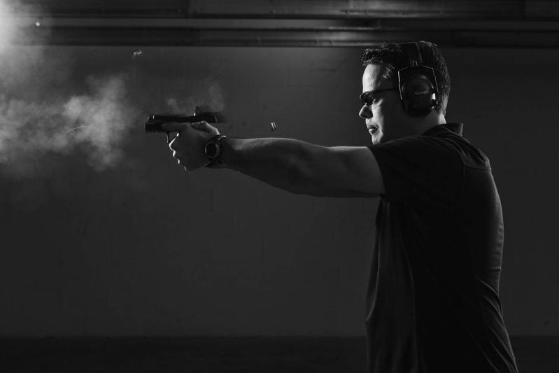 Ballistic team member firing a pistol in a dark indoor range with smoke and a spent casing in the air